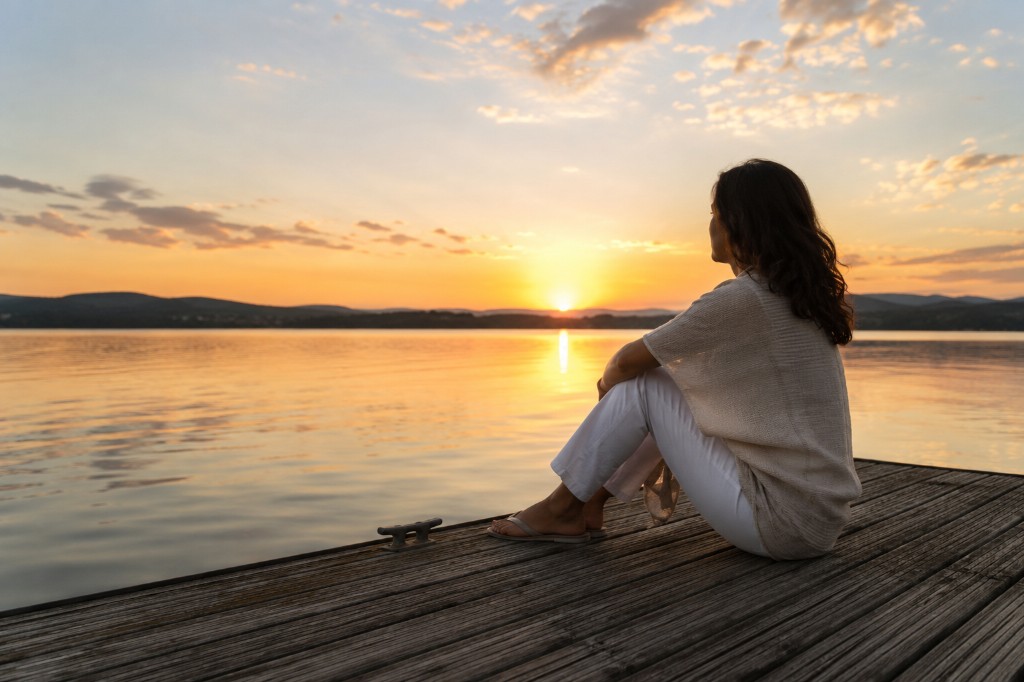 Woman sitting on a wooden dock at sunset, looking out over calm water